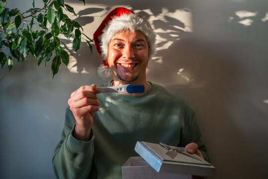 Man wearing a santa hat smiling happily, holding a positive pregnancy test and an open gift box, announcing a christmas surprise of impending fatherhood
