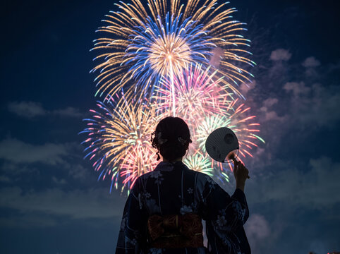 手裏剣の形の団扇を手に夜空の彩る大輪を見つめる着物姿の女性