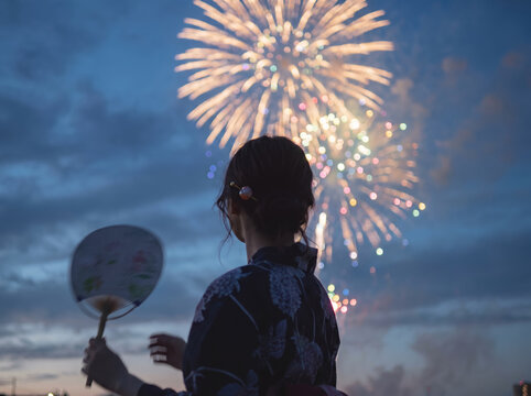 浴衣を着てうちわを持ち夜空の打ち上げ花火を見上げる女性