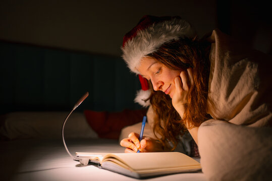Woman in santa hat writing christmas list or journaling notes in a notebook on bed with a small book lamp, feeling cozy and calm at night during holiday season, preparing for celebration