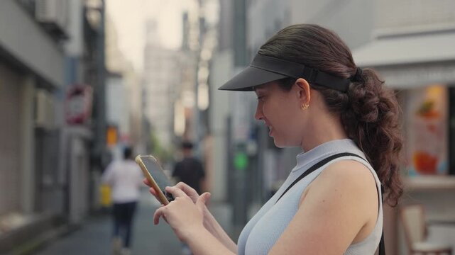 Happy caucasian woman wearing a sun visor using her smartphone for directions, texting, or browsing social media while standing on a narrow street during her vacation in Tokyo, Japan