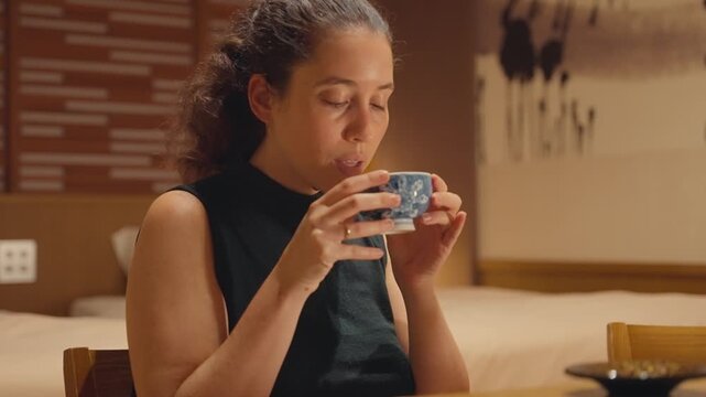 Beautiful caucasian woman relaxing and enjoying a hot cup of authentic Japanese green tea in a traditional hotel room, appreciating the aroma and flavour while closing her eyes