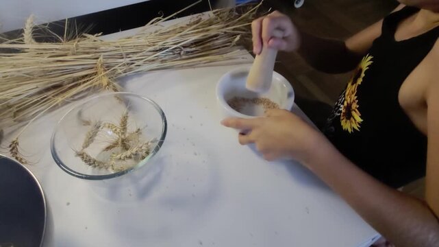 A high-angle wide shot shows a student steadying a ceramic mortar and crushing wheat grains with a wooden pestle beside bundles of harvested straw on a white table during a Waldorf farming lesson.