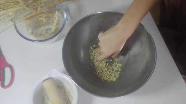 An overhead high-angle shot shows hands rubbing harvest wheat grains inside a large stainless steel bowl to separate chaff and husks from raw kernels during traditional post-harvest processing.