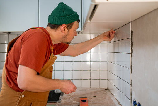 Man in work overalls and green beanie carefully applies grout spacers between white ceramic tiles on a kitchen wall, undertaking a renovation project and doing a home improvement task