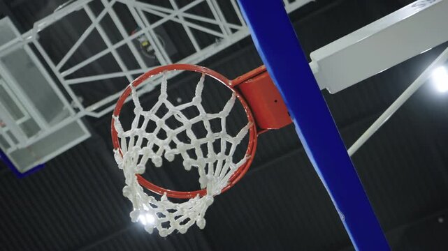 Basketball hoop net hangs inside brightly lit sports hall stadium. Red metal rim fastens securely to clear transparent glass backboard