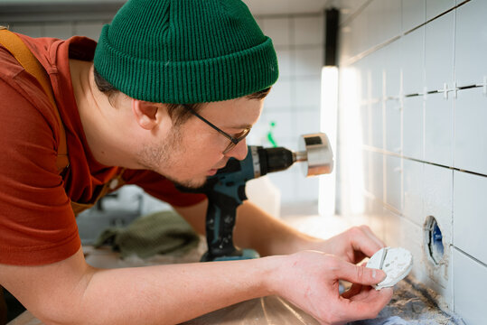 Man using a power drill with a core bit to cut a precise hole in ceramic kitchen wall tile for installing an electrical outlet during a home renovation project