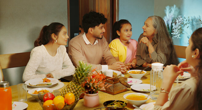 Indian happy relax joint family group sitting around table eat food talk speak share gossip have fun joy Young adult male female aged mom girl boy child spend time enjoy night dinner room home place