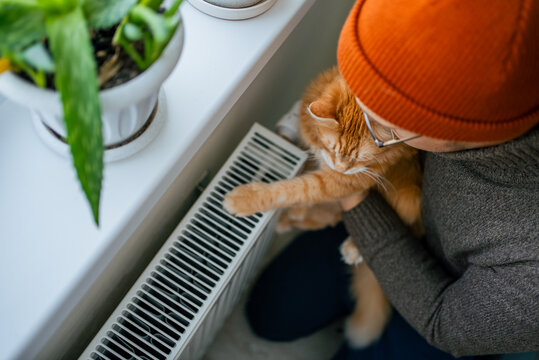 Person wearing an orange beanie is holding a sleeping ginger cat close to a warm radiator, symbolizing comfort and the act of reducing home heating expenses during the cold winter months