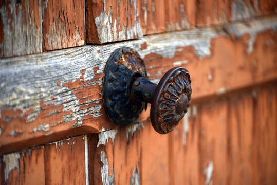 Close up of an old rusty door handle attached to a weathered brown wooden door with peeling paint, showing a rustic architectural detail full of character.
