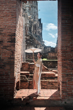 Portrait of Asian teenage girl dressed in traditional Thai outfit and umbrella with cheerful smile at old brick wall, summer holiday travel in Ayutthaya, ancient ruins heritage,  tourist destination.