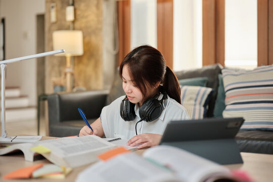 Asian teenage girl in headphones concentrates on learning at home, reading textbooks, note writing, self-tutorial exams, preparation for back to school, Homeschooling, university education lifestyle.
