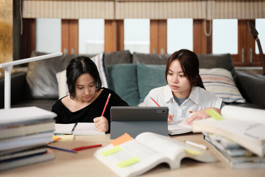 Two Asian teenage girls concentrate on online class, remote learning at home using tablet, writing and reading textbooks, self-study for back to school, Homeschooling, university education lifestyle.