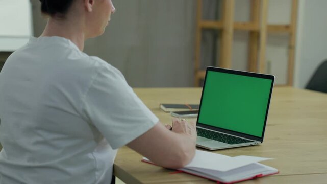 Woman Working On Laptop Computer with Green Mock Up Screen At Home Office Space. Female Freelancer Browsing Green Laptop Screen Writing Information in Notebook