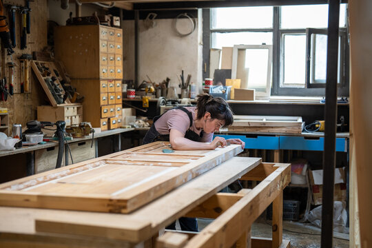 Passionate woman restorer carefully works on renewing old wooden door in creative atmosphere of restoration workshop, female crafter repairing details and elements with chisel, reconstruction process.