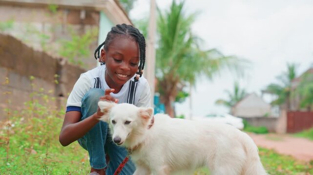 Black girl kneeling with white dog sunlit rural yard, palm trees and concrete wall, casual clothes, laughing while gently petting and coaxing dog, candid lifestyle moment with vibrant colors