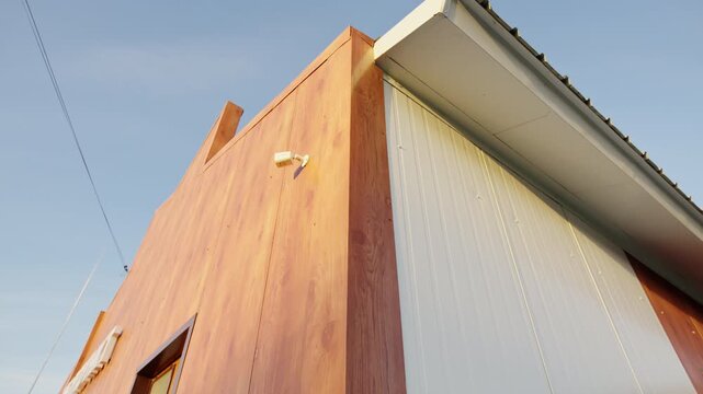 Compact security camera mounted on corner of brown wooden facade. Minimalist architectural details look modern against calm daytime sky