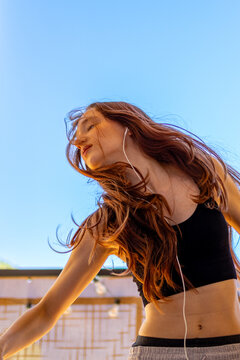 Red haired teenage girl dances passionately while listening to music through headphones on sunny balcony. Concept of energetic self expression and joyful dance movement outdoors with music.