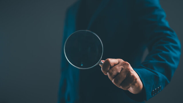 A person in a blue suit holds a magnifying glass, focusing on the viewer against a dark background.