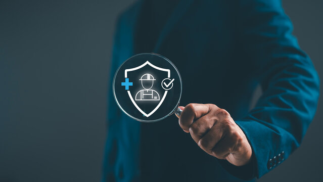 A person in a suit holds a magnifying glass focusing on a shield icon with a worker silhouette and medical cross, symbolizing workplace safety and health protection.