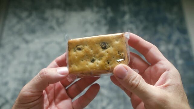 POV two hands holding small transparent package of crackers with raisins, inspecting healthy, convenient single serving snack against blurred background. Hands holding packaged crackers with raisins
