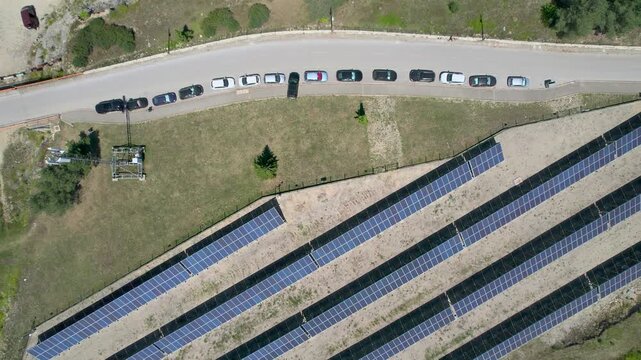 A top-down drone crosses parallel rows of solar panels and a lakeside road at Zaovine Lake in Serbia, with parked cars, rocky ground, forest edge and a small beach entering view.