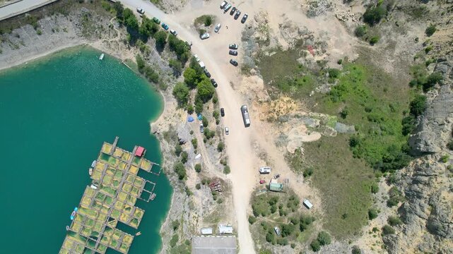 A top-down drone crosses floating fish pens at Zaovine Lake in Serbia, revealing a rocky shoreline, parked cars, dirt access road, small structures and green vegetation in daylight.
