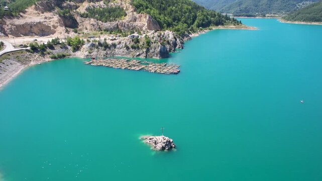 An aerial drone approaches floating fish pens along the rocky shore of Zaovine Lake in Serbia, passing a small islet as a dam road, quarry and dense forest fill the background.