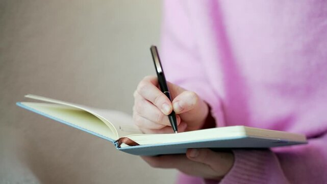 Close up of a woman writing in a notebook with a pen cozy at home. Female hands record notes, plans or personal thoughts, journaling, productivity, and everyday planning. Cropped.