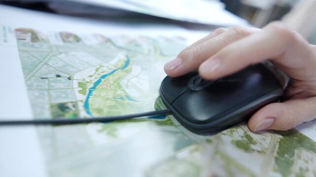 Human hand uses black computer mouse on top map. Wired optic device moves smoothly over detailed colorful urban plan during working process