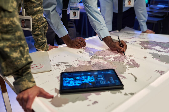 Diverse military officers and government officials analyzing world map on table during strategic planning session, suitable for national security and defense industry marketing