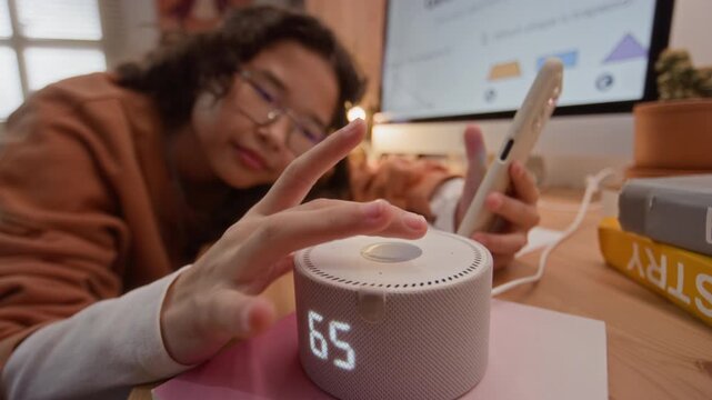 Asian teenager tapping smart speaker and holding smartphone while studying geometry online. Ideal for marketing e-learning platforms, educational apps, and smart home devices