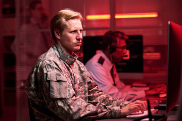Fototapeta premium Caucasian young adult soldier in camouflage uniform typing on computer keyboard in dark control room with red light. Image represents cybersecurity and military defense operations