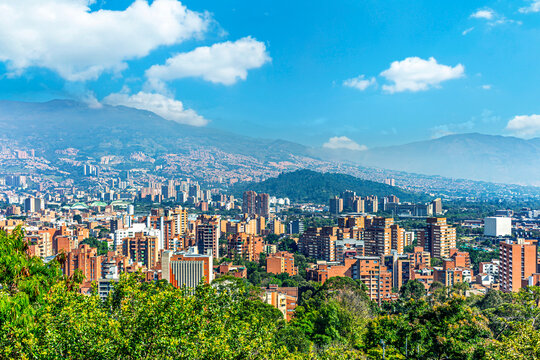 Panoramic view of the Medellin cityscape in Colombia. Red brick buildings and lush trees fill the valley, surrounded by majestic mountains under a bright blue sky with white clouds.