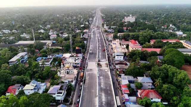 Aerial highlights modern road development in Kerala's scenic coastal region. Alappuzha Highway passing through Ambalappuzha with an under-construction overpass, moving vehicles, colorful neighborhood