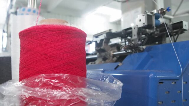 Close up shows red yarn spool feeding thread into industrial knitting machine. Automated textile equipment produces fabric inside modern factory.