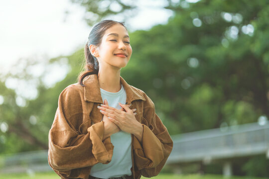 Young woman standing at park outdoors with hands over heart and eyes closed, expressing gratitude, mindfulness and inner peace