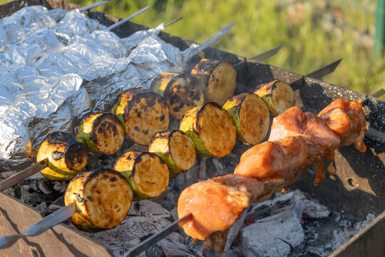 Delicious shish kebab and zucchini grilling on skewers over hot coals in a backyard grill. Meat and vegetables cooking outdoors during a sunny weekend picnic, preparing a tasty meal for family