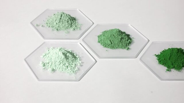 Vertical tracking shot of piles of different color powders in green shades on hexagonal watch glass pieces on a light grey backdrop lit with orbiting light and casts dynamic shadows
