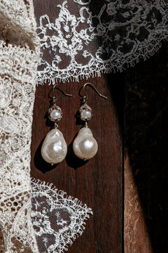 Elegant pearl drop earrings rest on dark wood beside delicate white lace, illuminated by dramatic directional sunlight.