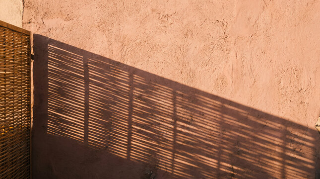 Woven bamboo fence casting striped sunlight shadow on pink terracotta stucco wall in minimalist boho style with copy space