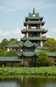 A majestic multi-tiered pagoda featuring green tiled roofs and red pillars stands beside a pond filled with lily pads in GuiHu Park, Chengdu. The scene captures a tranquil atmosphere under a clear