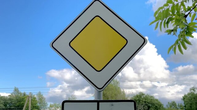 Priority road sign with yellow diamond on blue sky with white clouds and green tree leaves outdoors. Traffic symbol, main road, summer day.