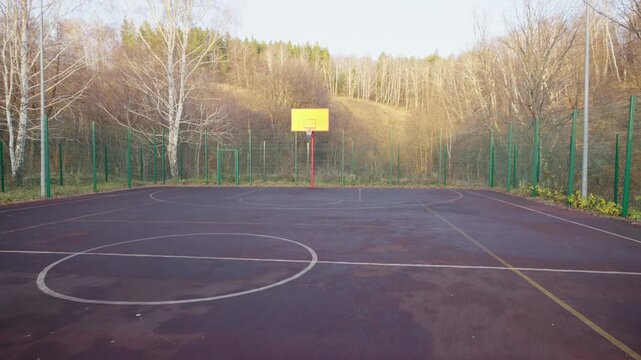 Empty basketball court occupies spacious fenced playground area. Golden autumn forest covers high hill behind court. Resort activity