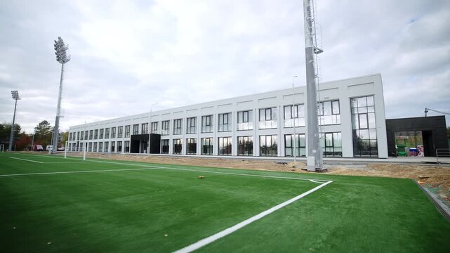 Contemporary white educational building stands next green soccer field. Sharp corner lines mark synthetic turf pitch surface clearly