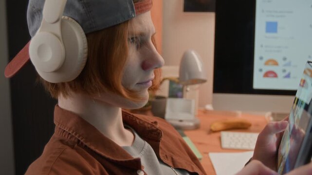 Caucasian teenager wearing headphones using tablet computer at desk with computer monitor in background. Clip is ideal for promoting online education, e-learning platforms, or digital lifestyle