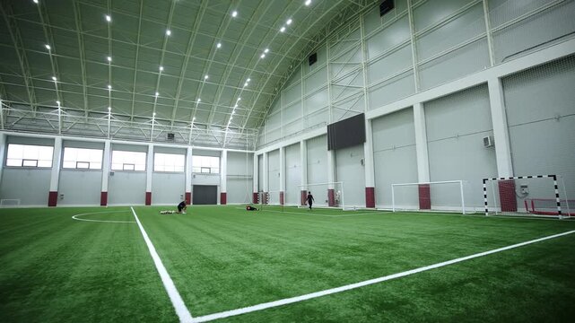 Players train on green artificial turf inside massive indoor arena. Bright spotlights under arched ceiling create focused sport mood