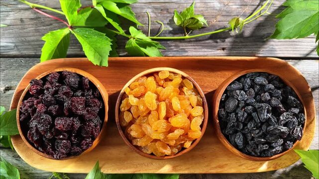 Horizontal overhead view of three raisin varieties in wooden bowls on a serving tray with grapevine leaves and downward movement