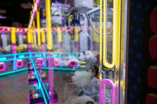Close-up captures the vivid neon framing in yellow, pink, and cyan that outlines the claw machine's glass and structure; blurred background reveals stacked plush prizes and mechanical components, emph
