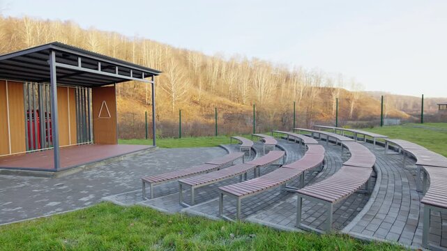 Outdoor theater area features curved wooden benches on paved semi-circle. Covered stage stands ready for performances on grassy hill slope